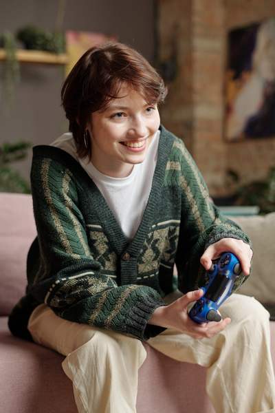 A girl with a pixie haircut in an oversize sweatshirt is smiling sitting on a light-colored sofa and playing a video game with a blue joystick